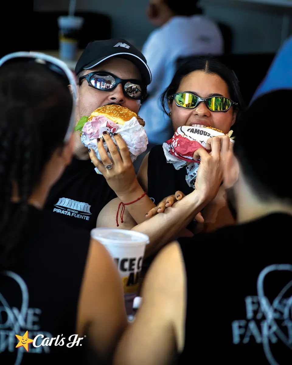 Dos corredores con gorra y lentes de sol muerden hamburguesas al mismo tiempo, todavía con la ropa de correr.