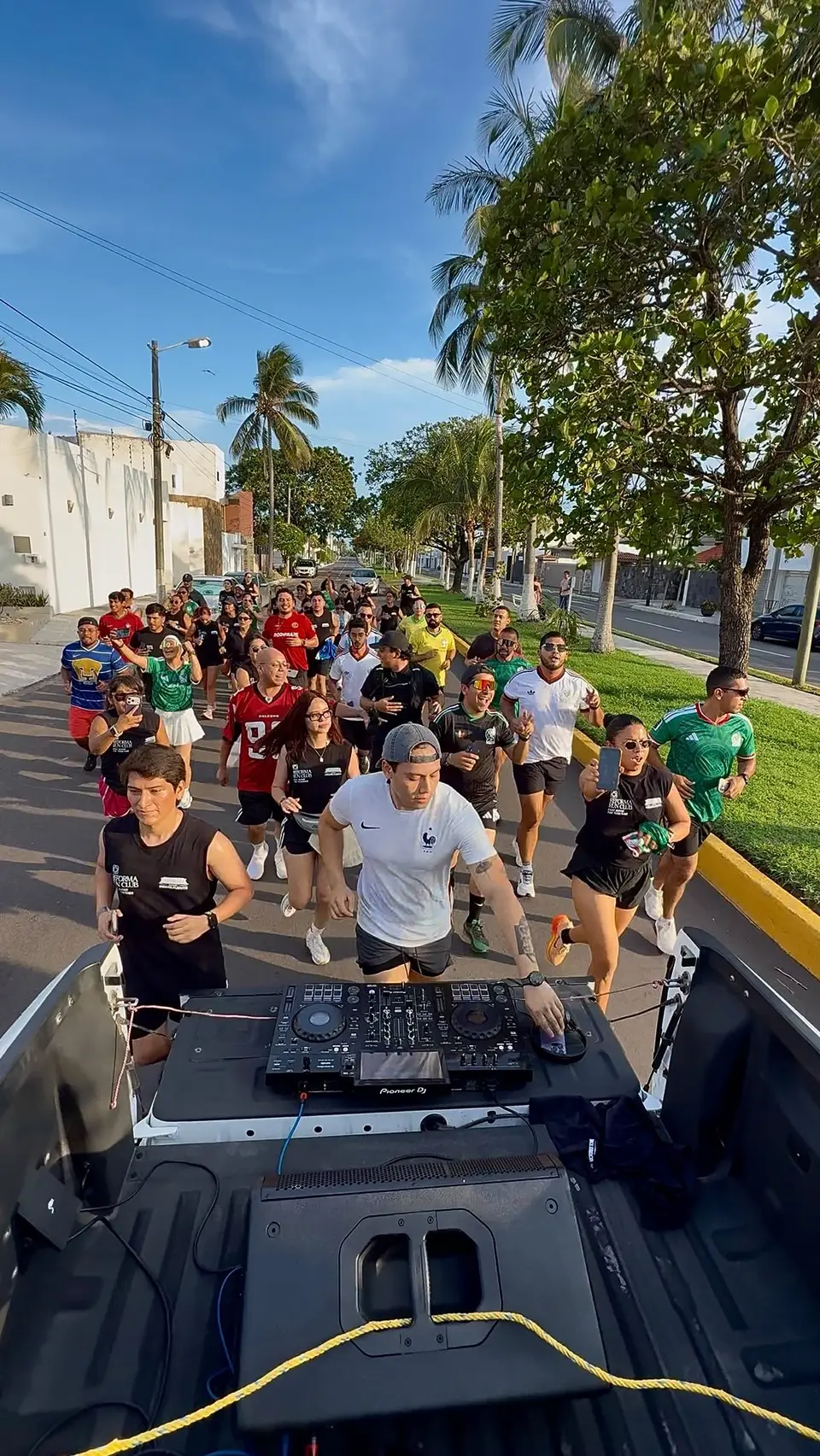 Un pelotón numeroso corre por la avenida bajo las palmeras, con la mesa de mezclas del DJ en primer plano desde la caja de una camioneta.